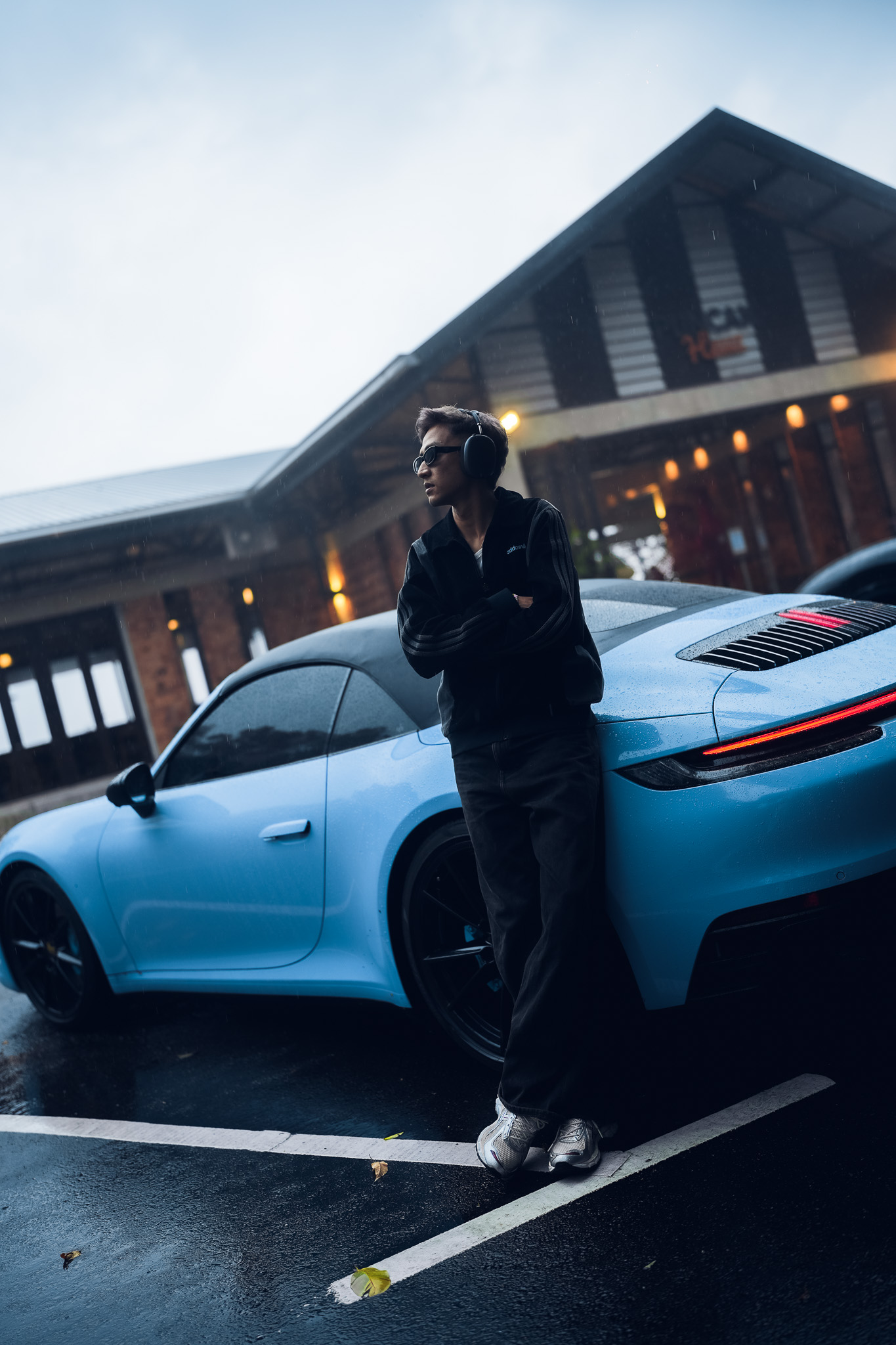 A person wearing headphones and sunglasses stands beside a blue sports car on a rainy evening.